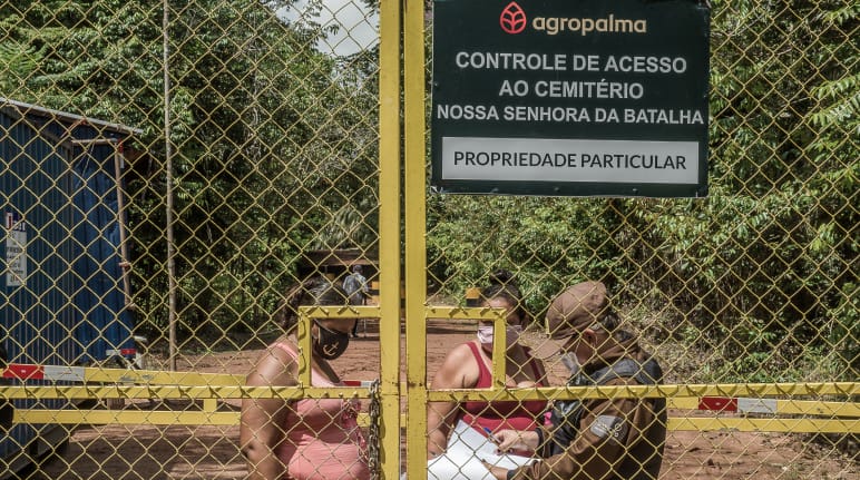 Deux femmes derrière une haute porte grillagée sont contrôlées par un garde de la société Agropalma. Le panneau de l’entreprise porte l’inscription : "Agropalma. Contrôle de l’accès au cimetière Nossa Senhora da Batalha. Propriété privée"