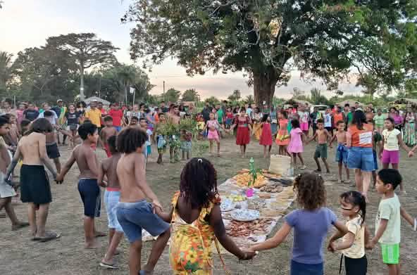 Des dizaines de participants, parmi lesquelles des femmes, des hommes, des enfants et des personnes âgées, forment un cercle autour d’une offrande posée à même le sol, composée d’aliments, de graines et d’autres produits de la terre