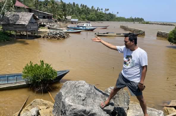 Amien Muhammad (Alliansi Sulawesi) montre l’eau polluée visible derrière lui, le long d’une côte tropicale.