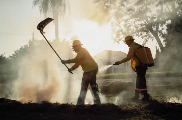 Deux hommes munis d’un batteur à feu et d’un sac à dos d’eau avec une pompe manuelle se trouvent dans la fumée.