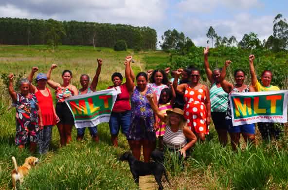 Un groupe de 15 femmes manifestent, le poing levé, avec deux banderoles sur lesquelles figurent les lettres « MLT »