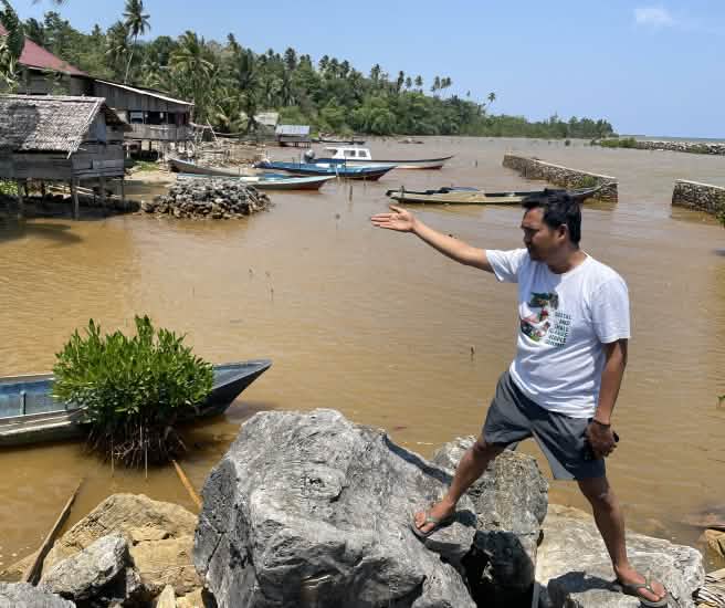 Amien Muhammad (Alliansi Sulawesi) montre l’eau polluée visible derrière lui, le long d’une côte tropicale.