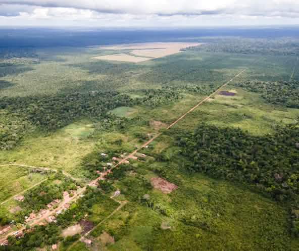 Vue aérienne d’une petite localité le long d’une piste rectiligne, avec derrière les plantations de palmiers à huile taillées dans la forêt tropicale