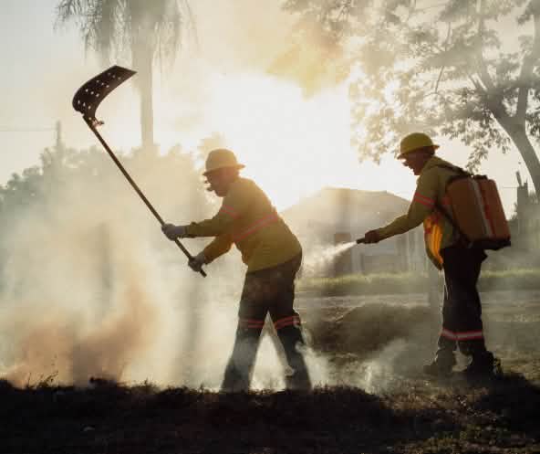 Deux hommes munis d’un batteur à feu et d’un sac à dos d’eau avec une pompe manuelle se trouvent dans la fumée.