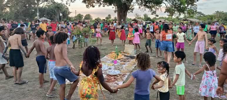 Des dizaines de participants, parmi lesquelles des femmes, des hommes, des enfants et des personnes âgées, forment un cercle autour d’une offrande posée à même le sol, composée d’aliments, de graines et d’autres produits de la terre