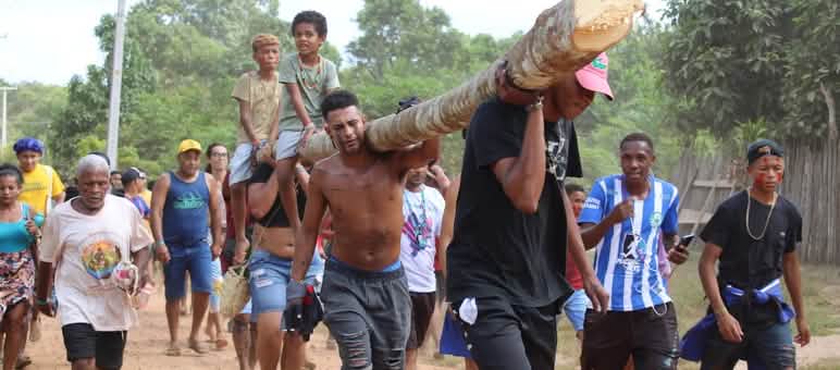 Des hommes et des femmes portent un tronc d’arbre sur leurs épaules. Deux enfants sont assis sur le tronc.