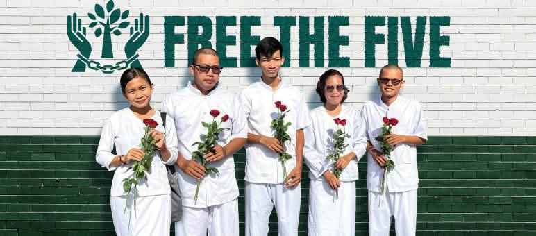 Cinq personnes vêtues de blanc, tenant des roses rouges, se tiennent devant un mur sur lequel on peut lire « Libérez les Cinq »