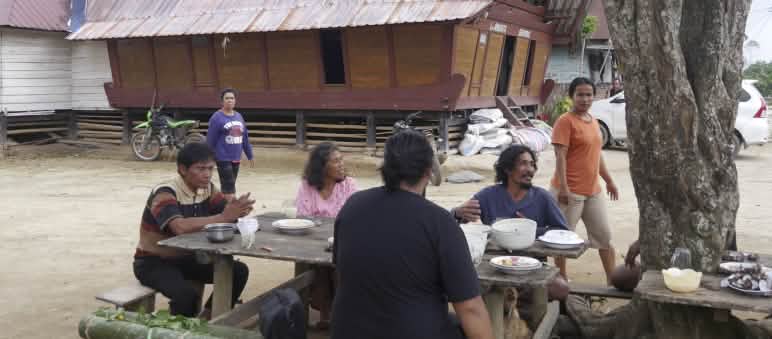 4 personnes assises à une table près d’un arbre, avec une maison batak en arrière-plan