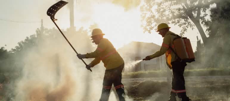 Deux hommes munis d’un batteur à feu et d’un sac à dos d’eau avec une pompe manuelle se trouvent dans la fumée.