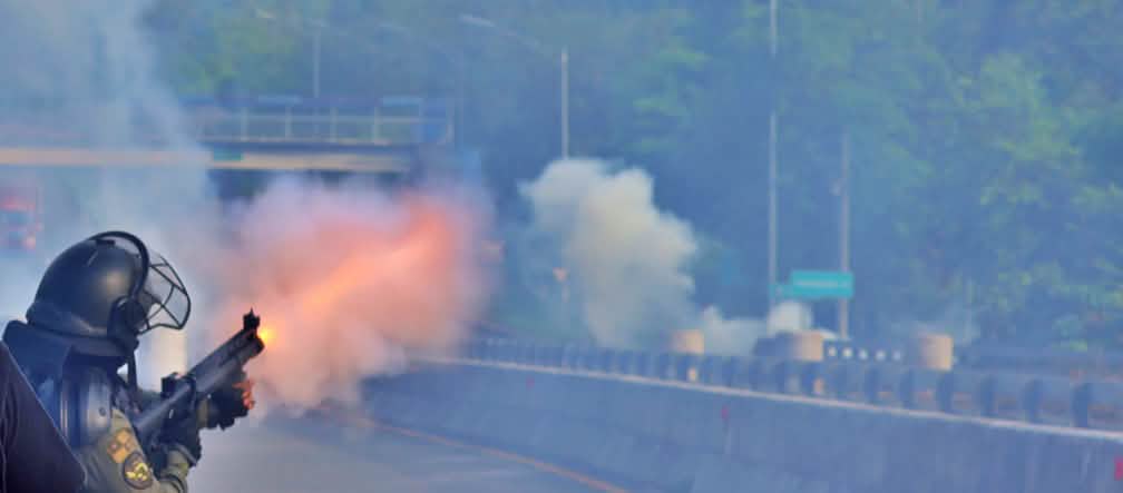 Un policier en tenue de protection noire tire des grenades lacrymogènes à l’aide d’un lanceur sur une autoroute déserte.