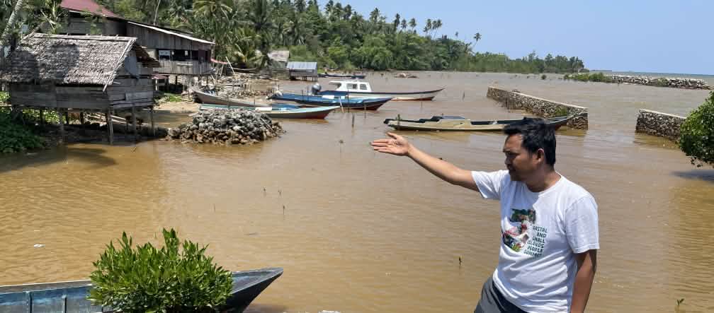 Amien Muhammad (Alliansi Sulawesi) montre l’eau polluée visible derrière lui, le long d’une côte tropicale.