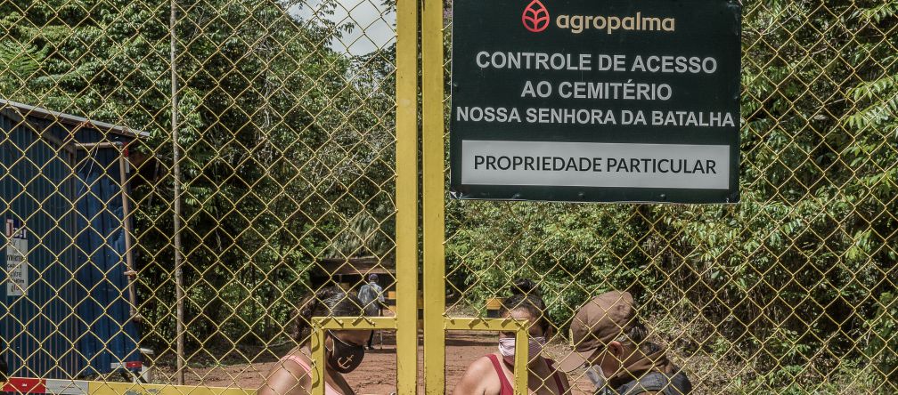 Deux femmes derrière une haute porte grillagée sont contrôlées par un garde de la société Agropalma. Le panneau de l’entreprise porte l’inscription : "Agropalma. Contrôle de l’accès au cimetière Nossa Senhora da Batalha. Propriété privée"