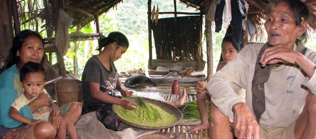 Dans une hutte ouverte, un homme, un petit garçon et trois femmes  sont assis, l’une d’elles trie du riz dans saladier plat