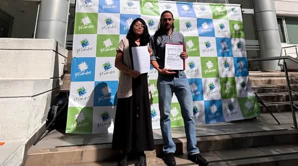 Une femme et un homme se tenant devant l’entrée d’un immeuble, à côté d’une banderole sur laquelle on peut lire « YASunidos », tenant des documents à la main