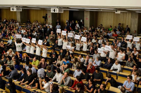 Vue plongeante sur l'amphithéâtre comble de l'université technique de Berlin. Debout, des activistes de Intag e.V. et Sauvons la forêt (Rettet den Regenwald) brandissent ensemble les pancartes floquées du nombre 61.431
