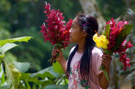 Une jeune fille Kichwa se tenant de profil porte un bouquet de fleur dans chaque main