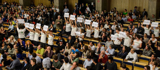 Vue plongeante sur l'amphithéâtre comble de l'université technique de Berlin. Debout, des activistes de Intag e.V. et Sauvons la forêt (Rettet den Regenwald) brandissent ensemble les pancartes floquées du nombre 61.431