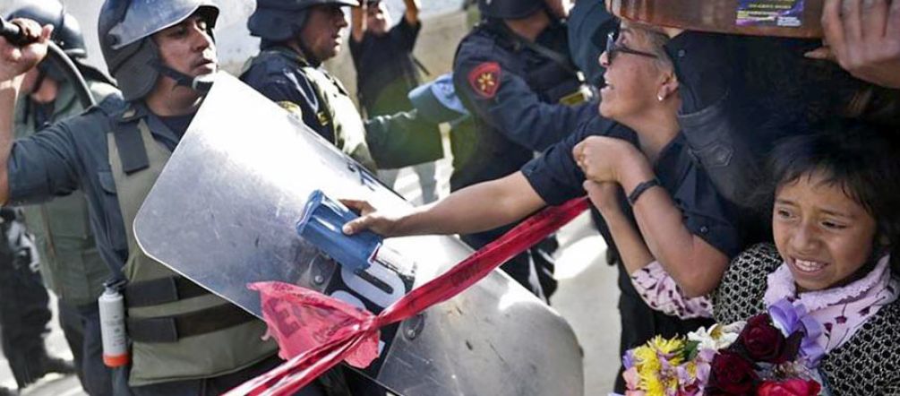 Violence policière contre les manifestants pacifiques à Cajamarca Un policier, protégé par son grand bouclier et son casque à visière, lève sa matraque contre une femme main dans la main avec une enfant au regard terrorisé qui tient un bouquet de fleurs