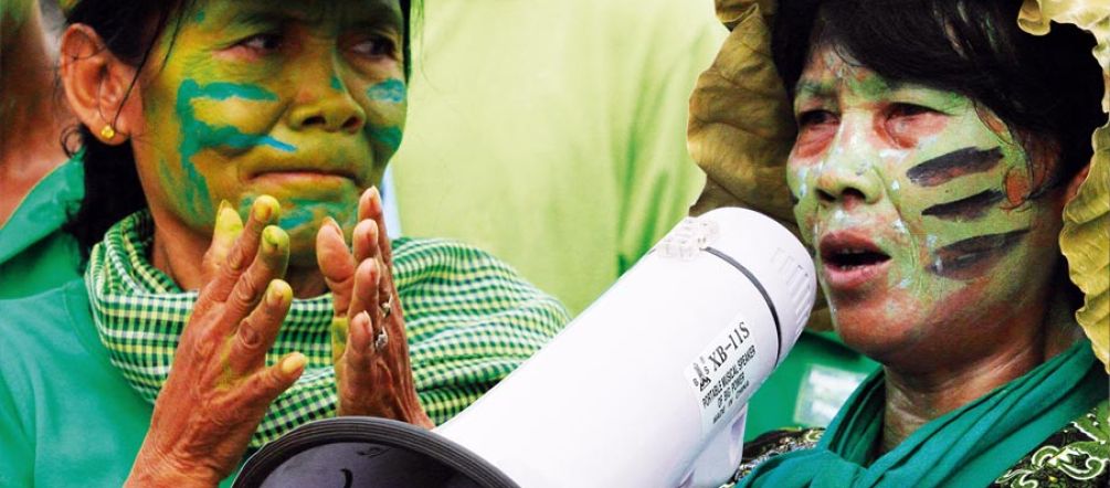 Gros plan sur deux femmes maquillées de bleu et de vert lors d'une manifestation. L'une tape dans les mains, l'autre parle dans un mégaphone