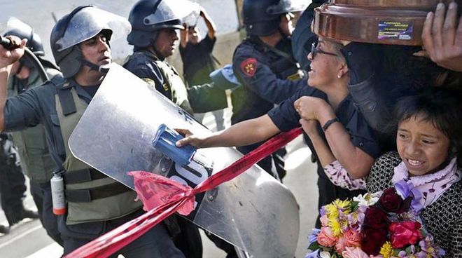 Violence policière contre les manifestants pacifiques à Cajamarca Un policier, protégé par son grand bouclier et son casque à visière, lève sa matraque contre une femme main dans la main avec une enfant au regard terrorisé qui tient un bouquet de fleurs