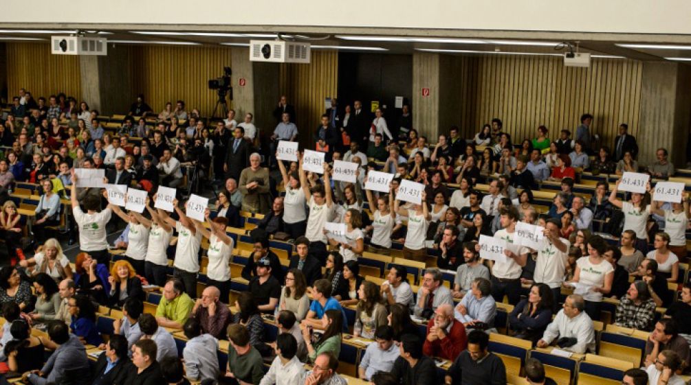 Vue plongeante sur l'amphithéâtre comble de l'université technique de Berlin. Debout, des activistes de Intag e.V. et Sauvons la forêt (Rettet den Regenwald) brandissent ensemble les pancartes floquées du nombre 61.431